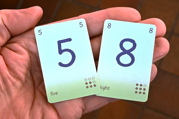 Two Dart Cards ~ a five and an eight ~ are held on an open hand to show the toned blue and green pastel background and the deepish blue and medium brown writing.